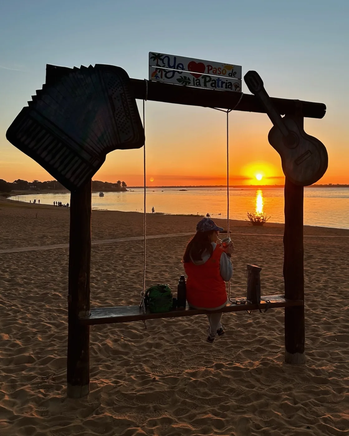 Persona tomando mate en el cartel Yo amo Paso de la Patria frente al atardecer
